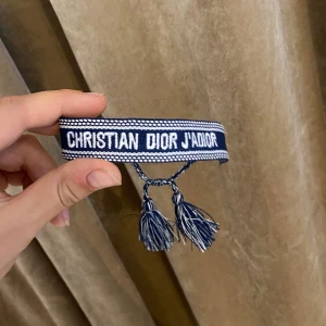 Blått armband Christian Dior J'Adior - Superfint blått armband från Christian Dior med texten 'J'Adior' broderad i vitt. Armbandet har snygga tofsar i ändarna och är gjort i ett mjukt tygmaterial. Perfekt accessoar för att lyfta din outfit med en lyxig touch.