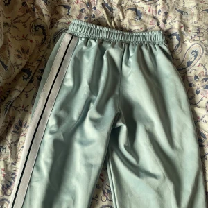 Palm Angels ljusblå mjukisbyxor - Palm Angels mjukisbyxor i ljusblått med vita och svarta ränder längs sidorna. Byxorna har elastisk midja med snörning och Palm Angels-logga tryckt på framsidan. Tillverkade i Italien, materialet är mjukt och har en snygg streetwear-vibe.