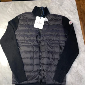 Svart Moncler cardigan - Snygg svart cardigan från Moncler med quiltad front och rygg i glansigt material och stickade ärmar samt krage. Jackan har dragkedja framtill, två fickor med dragkedja och Moncler-logga på ärmen. Perfekt för dig som vill ha en stilren och exklusiv look.