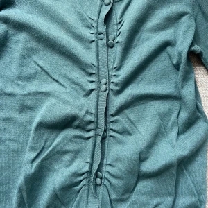 Grön kofta från Vero Moda med knappar - Söt, grön kofta från Vero Moda i storlek S. Koftan har dekorativa tygklädda knappar framtill och fina rynkade detaljer längs knappraden. 3/4 långa ärmar och i mjukt stickat material.