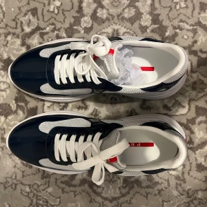 Prada sneakers i vitt och marinblått - Snygga Prada sneakers med vit mesh och marinblå lackdetaljer. Skorna har vit platt sula, rund tå och klassisk snörning. Röd Prada-logga på innersulan och yttersulan. Perfekt för dig som vill ha en clean och sportig look med lyxig känsla.