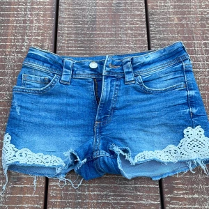Blå jeansshorts med spetsdetaljer - Snygga blå jeansshorts med råa kanter och vita spetsdetaljer längs bensluten. Klassisk femficksmodell med bälteshällor och dragkedja. Perfekta för varma dagar när du vill ha en chill och trendig look.
