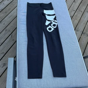 Svarta leggings från Adidas med logga - Svarta leggings från Adidas med stor vit logga längs ena benet. Tight passform och stretchigt material som passar perfekt till träning eller chill. Klassisk sportig look med hög midja och bekväm resår i midjan.