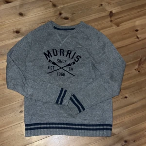 Grå stickad tröja från Morris - Snygg grå stickad tröja från Morris med mörkblå tryck och detaljer. Tröjan har rund hals, långa ärmar och ribbade muddar med blå ränder vid ärmslut och nederkant. Perfekt för sommarkvällarna