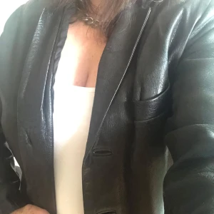 Svart skinnkavaj med knappar - Snygg svart kavaj i äkta skinn med klassisk krage och tre knappar framtill. Flera fickor och stilren passform, perfekt för dig som vill ha en edgy men stilren look. Fodrad insida och lång ärm. Passar till många olika outfits. Modellen är large. Inköpt på loppis i Amsterdam. Fodret kan behöva lite kärlek. 