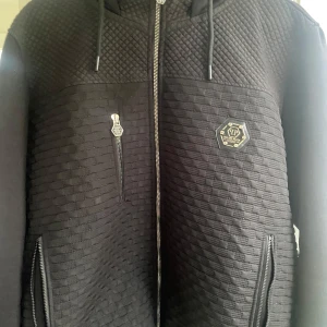 Svart Jacka/Hoodie från Philipp Plein - Säljer en svart Jacka/Hoodie från Philipp Plein med unik textur och mönstrad framsida. Jackan har dragkedja, huva med snörning och flera fickor med dragkedja. Insidan är fodrad med logotypmönstrat tyg. Perfekt för dig som vill sticka ut med stil.