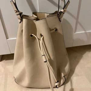 Beige bucket bag från H&M med snörning - Stilren beige bucket bag från H&M i syntetmaterial med justerbar axelrem och snygga guldfärgade metalldetaljer. Väskan har snörning upptill och kommer med en matchande liten innerpåse. Perfekt för dig som vill ha en clean och modern look.