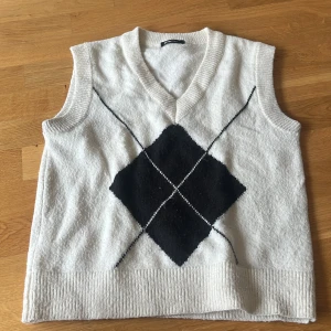 Vit stickad väst med argylemönster - Vit stickad väst med svart argylemönster framtill och v-ringad hals. Västen har bred ribbad kant nedtill och runt ärmhålen. Perfekt för lager-på-lager och ger en klassisk preppy vibe.