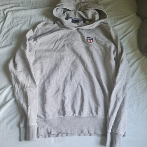 Ljusgrå hoodie från GANT med brodyr - Snygg ljusgrå hoodie från GANT med broderad logga på bröstet. Klassisk modell med huva och långa ärmar. Tillverkad i mjuk bomullsmix som känns skön mot huden. Perfekt för en chill och stilren look. Skriv vid intresse