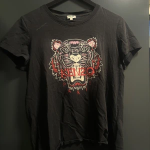 Svart Kenzo t-shirt  - Äkta Svart t-shirt från Kenzo med ikoniskt tigertryck och röd Kenzo Paris-text på bröstet. Klassisk passform och rund halsringning, tillverkad i mjuk bomull. Perfekt statement-plagg för dig som gillar streetstyle och vill sticka ut. Behöver tvättas därav priset nypris 1299kr