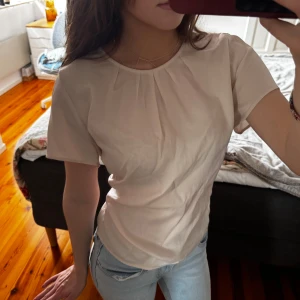 Ljusbeige blus med veckad hals - Säljer en ljusbeige blus med korta ärmar och rund halsringning. Blusen har fina veck framtill vid halsen som ger en snygg detalj. Den är tillverkad i ett tunt, lätt material som känns luftigt och skönt. Perfekt att styla med jeans eller kjol.