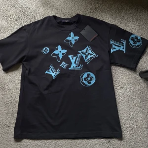 Svart Louis Vuitton t-shirt med blå logga - Snygg svart t-shirt från Louis Vuitton med stora blå LV-loggor och blommönster tryckta på bröstet och ena ärmen. Klassisk passform och rund halsringning, perfekt för dig som vill sticka ut med en exklusiv streetstyle-look.