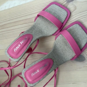 Rosa sandaler med klack från Public Desire - Trendiga sandaler med fyrkantig tå och klack i rosa från Public Desire. Skorna har flätad beige textil på sulan, rosa syntetdetaljer och långa remmar att knyta runt vristen. Perfekta för att lyfta din sommarlook.