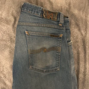 Ljusblå jeans från Nudie Jeans - Säljer ett par ljusblå jeans från Nudie Jeans med klassisk femficksdesign och snygga slitningar. Jeansen har kontrastsömmar och Nudie-logga på bakfickan. Perfekt för dig som gillar en avslappnad och trendig look.