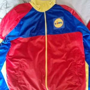 LIDL tracksuit i rött, blått och gult - Färgstark tracksuit från LIDL med kavajjacka och byxor i rött, blått och gult. Jackan har dragkedja och LIDL-logga på bröstet, medan byxorna har blå snörning och loggor längs sidorna. Tillverkad i mjuk polyester med sportig look. 229kr styck eller 400kr tillsammans