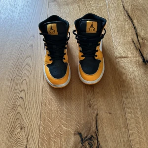 Nike Air Jordan 1 Mid svart/gul - Säljer ett par Nike Air Jordan 1 Mid i svart, gul och vitt. Klassisk high-top siluett med svart tå och snörning, gula paneler och vit sula. Ikonisk Swoosh på sidan och Jumpman-logga på plösen. Perfekta för dig som vill sticka ut med din sneakerstil.