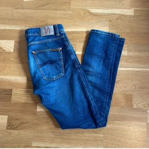 Blå jeans från Nudie Jeans - Säljer ett par klassiska blå jeans från Nudie Jeans med snygg tvätt och smal passform. Jeansen har fem fickor, orangea sömmar och Nudie-logga på bakfickan. Perfekta för dig som gillar en modern och stilren look.