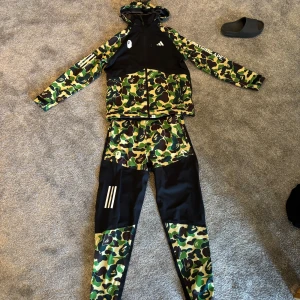 Adidas x BAPE camo träningsset - Säljer ett fett träningsset från Adidas och BAPE med ikoniskt grönt camouflage-mönster och svarta paneler. Setet består av en jacka med huva och dragkedja samt matchande byxor, båda med loggor och vita detaljer. Perfekt för dig som vill sticka ut på gymmet eller gatan.