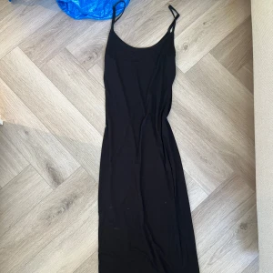 Svart långklänning med smala axelband - Enkel och stilren svart långklänning med smala axelband och rundad urringning. Klänningen har en tight passform och är perfekt för dig som gillar minimalistisk stil. Materialet känns mjukt och följsamt mot kroppen.