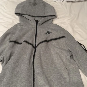Grå Nike hoodiejacka med dragkedja - Snygg grå hoodiejacka från Nike med hel dragkedja framtill, svart Nike-logga på bröstet och svarta detaljer längs bröstet och ärmen. Jackan har huva och en praktisk ficka med dragkedja på ärmen. Perfekt för en sportig och avslappnad stil.