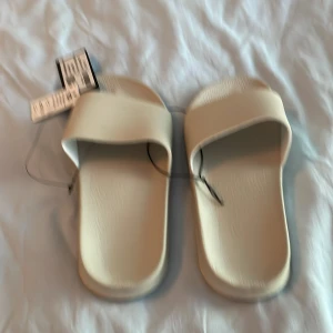 Beige badsandaler i syntet - Snygga och minimalistiska badsandaler i beige från 157. De har en bred rem över foten och en platt sula med mönstrad undersida för extra grepp. Perfekta att glida i till stranden eller poolen. Tillverkade i lätt och bekvämt syntetmaterial.