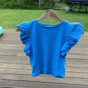 Blå ärmlös topp med volang - Supersöt blå topp i bomull med breda volangärmar som ger en extra cool touch. Modellen är ärmlös och har rund halsringning. Perfekt för dig som vill sticka ut med en färgstark och lekfull stil.                                                        Själv skulle jag säga att den är stl xxs-xs eller 135-140