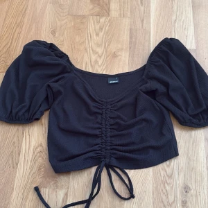 Svart croppad blus från Gina Tricot - Supersnygg svart croppad blus från Gina Tricot med puffiga korta ärmar och v-ringning. Toppen har trendig snörning framtill som ger en cool rynkad effekt. Perfekt för dig som gillar statement-ärmar och en feminin siluett.