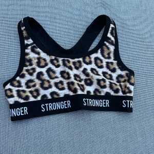 Leopardmönstrad sporttopp Stronger XS - Sportig leopardmönstrad topp från Stronger i storlek XS. Toppen har bred resår med logga, svarta detaljer och är ärmlös med klassisk sport-bh form. Perfekt för dig som vill sticka ut på gymmet eller under träningspasset.