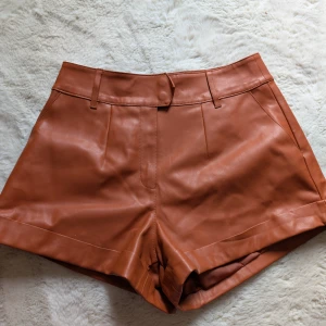 Bruna skinnshorts med hög midja - Oanvända Snygga bruna shorts i skinnimitation med hög midja. De har bälteshällor, diskreta fickor och stilrena sömmar framtill. Perfekta för dig som vill ha en trendig och edgy look till sommaren.