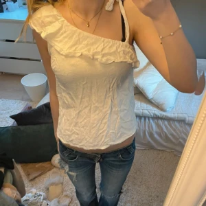 Vit omlottopp med volang och knyt - Säljer en söt vit topp med omlottkänsla och volang längs den asymmetriska halsringningen. Toppen har en axelbandslös sida och ett axelband med knyt på andra sidan. Perfekt för varma dagar och ger en fräsch look.