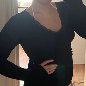 Svart långärmad topp med spets - Svart långärmad topp med v-ringning och snygga spetsdetaljer runt halsen. Tight passform som framhäver figuren och ger en stilren look. Perfekt att styla med jeans eller kjol för en trendig outfit.
