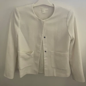 Vit kavajjacka - Stilren vit kavajjacka från H&M med rund halsringning och två stora fickor framtill. Jackan har diskreta knappar och är tillverkad i ett strukturerat tyg. Perfekt klassisk och trendig sommarjacka! 