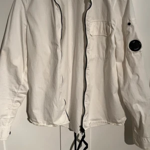 Vit overshirt från C.P. Company - Snygg vit overshirt från C.P. Company med klassisk linsdetalj på ärmen. Jackan har dragkedja framtill, bröstficka och knappar vid ärmslut. Tillverkad i bomull, perfekt för lager-på-lager och stilrena outfits.