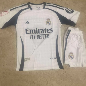 Real Madrid Bellingham matchtröja set - Real Madrid fotbollströja och shorts med Bellingham 5-tryck. Vit med mörkblå detaljer, klubbmärke och Adidas-logga. Kortärmad modell i lätt funktionsmaterial, perfekt för fotbollsträning eller match.