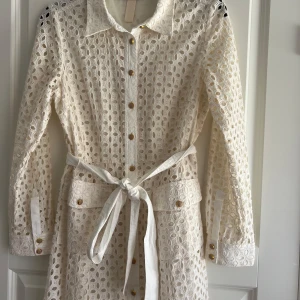 Vit spetsklänning med bälte från Zara - Superfin vit spetsklänning från Zara med lång ärm, snygga guldknappar och knytbälte i midjan. Klänningen har ett genombrutet broderat mönster och stora fickor framtill. Perfekt för dig som gillar romantisk och trendig stil.