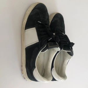 Valentino Garavani svarta sneakers med nitar - Svarta och vita sneakers från Valentino Garavani med coola silverfärgade nitar på hälen. Ovandel i mocka och skinn, klassisk rund tå och snörning. Snygg kontrast mellan svart mocka och vita skinnpaneler. Perfekta för dig som vill sticka ut med en lyxig streetstyle. En nit saknas på varje häl. 