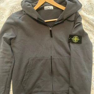 Grå hoodie från Stone Island - Säljer en grå hoodie från Stone Island med dragkedja och klassisk logga på ärmen. Tröjan har huva, känguruficka och är tillverkad i mjuk bomull. Perfekt för dig som gillar streetwear och vill ha en stilren look.