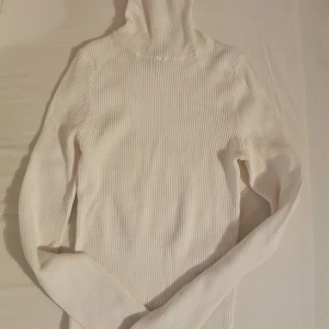 Vit ribbad polotröja - Snygg vit polotröja med ribbad struktur och lång ärm. Perfekt att styla med jeans eller kjol för en clean och fräsch look. Hög krage som ger extra stilpoäng och håller dig varm under kyliga dagar.
