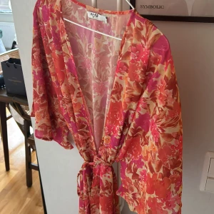 Blommig omlottklänning från NA-KD - Färgstark omlottklänning från NA-KD med blommigt mönster i rosa, rött och orange toner. Klänningen har v-ringning, vida ärmar och knyt i midjan för en snygg siluett. 