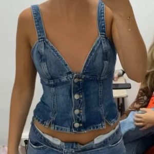 Jeanskorsett med knappar - Säljer en croppad jeanskorsett i klassisk blå denim med breda axelband och snygg v-ringning. Toppen har markerade sömmar och knäppning framtill med fyra silvriga knappar. Perfekt för dig som vill ha en edgy och trendig look.