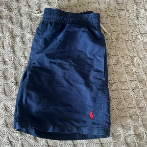 Marinblå badshorts från Polo Ralph Lauren - Snygga marinblå badshorts från Polo Ralph Lauren med röd broderad logga framtill. Elastisk midja med snörning och innerbyxa i mesh. Klassisk passform och bakficka med kardborre. Perfekta för stranden eller poolen.