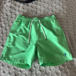 Gröna badshorts Polo Ralph Lauren - Snygga gröna badshorts från Polo Ralph Lauren med vit snörning i midjan och blå logga broderad på benet. Shortsen har elastisk midja, sidofickor och en bakficka med knapp. Tillverkade i lätt polyester med meshfoder, perfekta för stranden eller poolen.