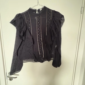 Svart blus med paljettdetaljer S - Supersnygg svart blus med transparenta partier, broderade blommor och glittriga paljettränder framtill. Långa ärmar med spets och volangdetaljer ger en romantisk vibe. Perfekt för dig som gillar unika detaljer och en lite lyxigare känsla.
