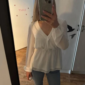 Vit omlottblus med v-ringning - Säljer en luftig vit blus med omlottdesign och v-ringning. Blusen har långa, transparenta ärmar med resår vid handlederna och markerad midja för en snygg siluett. Perfekt att styla med jeans eller kjol för en fräsch look.