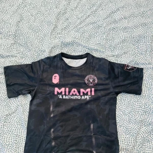 Inter Miami x BAPE fotbollströja svart - Snygg svart Inter Miami fotbollströja i samarbete med BAPE. Tröjan har rosa tryck med MIAMI och BAPE-logga på bröstet samt klubbmärke. Kortärmad modell i lätt och sportigt material, diskret camo-mönster i tyget och tryck på ena ärmen.