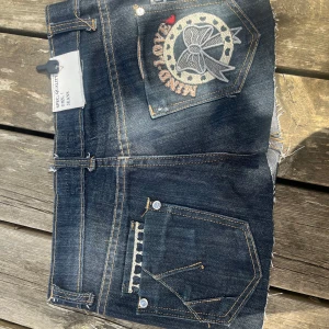 Lågmidjad jeanskjol - Snygg mörkblå jeanskjol  med råa kanter och cool broderad bakficka med texten 'KIND LOVE' och en stor rosett. Klassisk femficksmodell, kontrastsömmar och knapp med logga. Perfekta för sommaren och ger en edgy vibe.