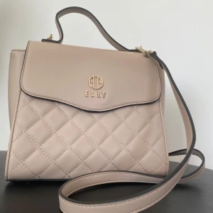 Beige quiltad axelväska från Elly - Snygg beige axelväska från Elly med quiltad framsida och guldfärgade detaljer. Väskan har både handtag och justerbar axelrem, samt dragkedja och magnetlås. Insidan har flera fack för smart förvaring. Perfekt för dig som vill ha en stilren och trendig väska.