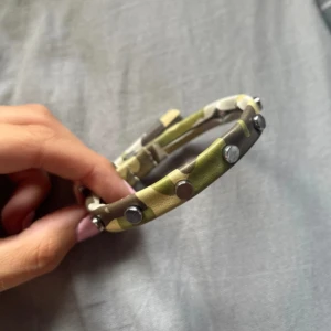 Armband med nitar och spänne - Snyggt armband i camouflage-mönstrat konstläder med silverfärgade nitar och klassiskt spänne. Färgerna går i grönt, beige och brunt, vilket ger en cool och edgy vibe. ☺️