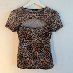 Leopardmönstrad topp  - Snygg leopardmönstrad topp från SHEIN i brun och svart med korta ärmar och cool cut-out detalj vid bröstet. Toppens material är stretchigt och tunt, perfekt för att sticka ut. Passformen är tight och den har rund halsringning. Storlek S