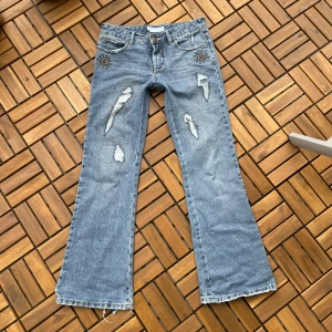 Bootcut jeans med slitningar och nitar - Ljusblå bootcut jeans med slitna detaljer och råa kanter. Framtill finns coola nitar vid fickorna och flera slitningar för en edgy look. Bakfickorna har broderade detaljer. Jeansen är i klassisk denim och har en avslappnad passform. Defekter finns på bilderna! Mått: midjemåttet tvärsöver 39cm, Innerbenslängden 74.5-75 cm. Storleken är 14år men skulle säga att de är storlek S💗 Kolla gärna in allt annat jag säljer!🥰